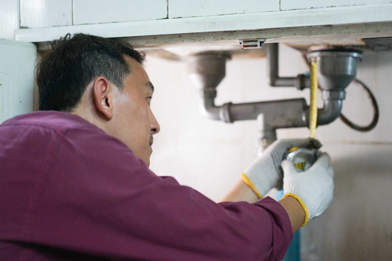 A male plumber repairs a pipeline or drain under the sink in kitchen room.