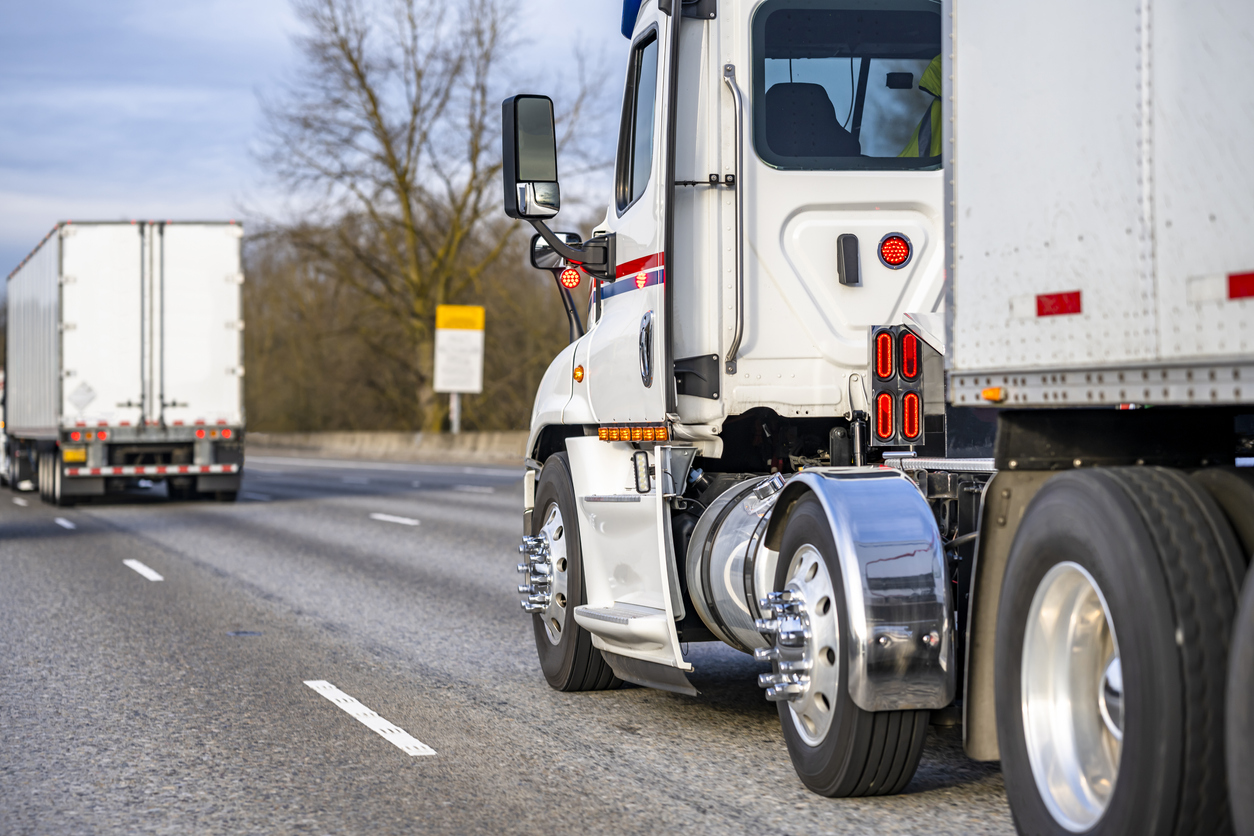 Day cab white big rig semi truck with chrome fenders transporting cargo in dry van semi trailer running in convoy behind another semi truck driving on the interstate highway road